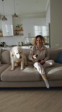 Woman Sitting On Bright Living Room Sofa Reading Messages On Smartphone When Golden Retriever Jumps Over Sofa Back Holding TV Remote And She Smiles Taking It Turning On Television Creating Warm