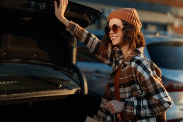 Woman with beanie and sunglasses smiling while loading shopping into a car trunk at golden hour, candid lifestyle moment in plaid coat with authenticity, mindful living and emotional storytelling.