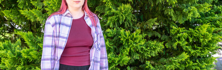 young Caucasian woman with bright pink hair stands in front of a green arborvitae hedge. She is wearing a plaid shirt and a burgundy top. Copy space