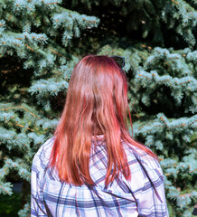 young woman with long, red brown hair stands with her back to the camera, surrounded by green spruce trees. She wears a plaid shirt
