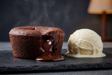 A warm, steaming chocolate lava cake with a flowing molten center is served next to a scoop of vanilla ice cream on a rustic slate board. 