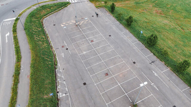 Overhead parking lot view showing lone cyclist crossing white lined area beside verdant grass and roadside stop sign