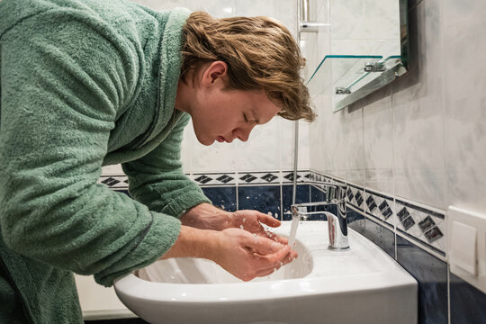 Young man in bathrobe washing his face with water over bathroom sink as part of his morning hygiene and skincare routine. Side view. - Powered by Adobe