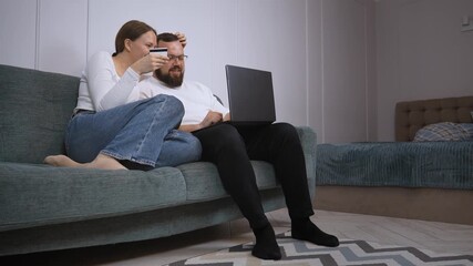 Young couple relaxing on a comfortable sofa at home, using a laptop for online shopping and holding a credit card, enjoying a convenient and modern retail experience - Powered by Adobe