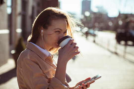 Young adult woman smiling, sipping coffee on sunny city street