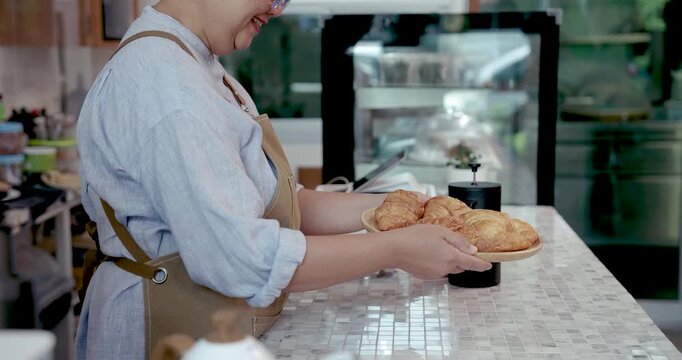 Asian senior female barista smiling while holding plate of fresh croissants behind coffee shop counter showing hospitality and service in cozy small retirement business