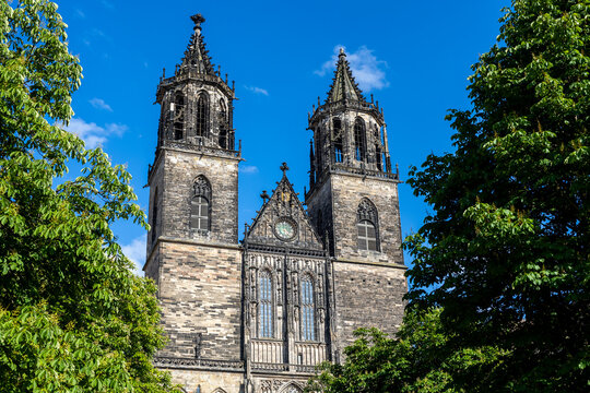 Majestic Magdeburg Dom towers rise above lush green trees under vibrant blue sky. Gothic stone details, pointed spires and central clock create striking contrast with bright summer atmosphere - Powered by Adobe