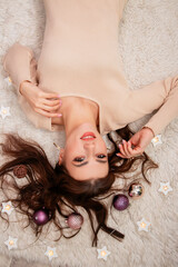 Top view portrait of young woman lying on fur rug surrounded by Christmas balls and garlands wearing beige tight knitted dress