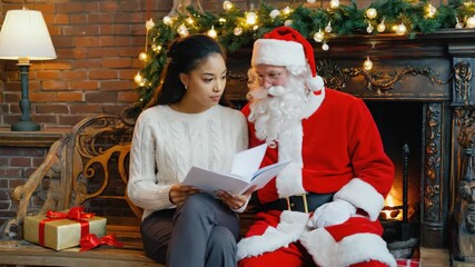 Young mixed race woman and santa claus reading a storybook together in a festive home. Celebrating christmas traditions with a magical moment of storytelling by the fireplace - Powered by Adobe