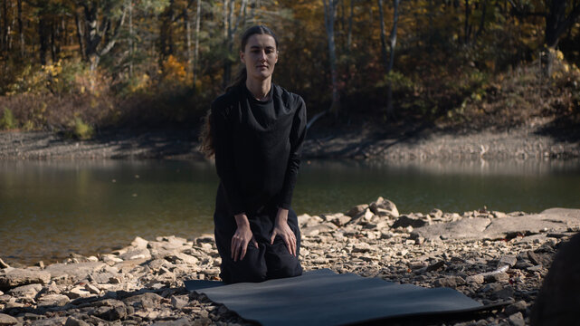 Woman kneeling on rocky riverside with yoga mat in nature - Powered by Adobe