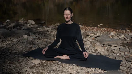 Fotobehang Lotusbloem Woman meditating in lotus pose on yoga mat by rocky riverbank outdoors  © Mariia