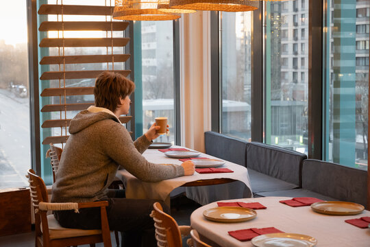Young caucasian man sitting alone at restaurant table with city view, holding paper coffee cup and relaxing in quiet interior. Coffee Break at cafe