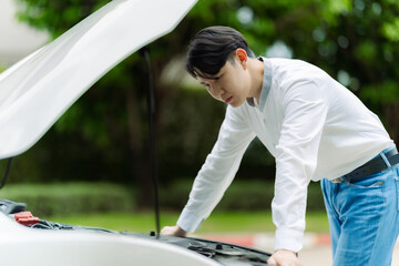 Young Asian man checking the engine of a broken down car parked on the side of a country road....