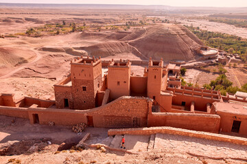 A person is walking down stone steps outside the Ksar of Ait ben haddou, a fortified city in Morocco. The desert stretches out beyond the walls.