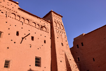 The earthen buildings of the historic Ksar of Ait ben Haddou rise against a clear blue sky. The traditional architecture shows detail and texture.