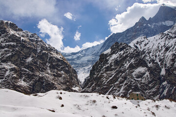 A snowy path winds through the high altitude Annapurna region of Nepal. Rocky mountains tower on either side, leading towards Annapurna Base Camp, under a blue sky