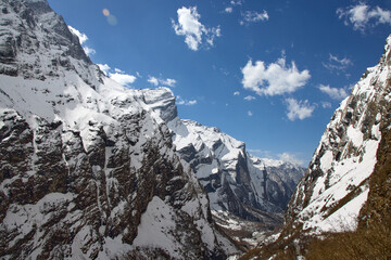 This captures the trail to Annapurna Base Camp in Nepal. Majestic Himalayan mountains, covered in snow, rise on either side under a bright blue sky with scattered clouds.