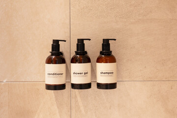 Brown bottle mockup for bathing products in hotel bathroom, spa shampoo, shower gel, liquid soap, body wash, and conditioner dispensers on shower wall.