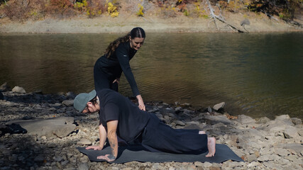Outdoor yoga instructor assisting person in plank position by riverside