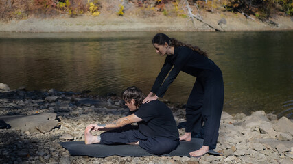 Woman assisting man with yoga stretching beside a river on rocky ground