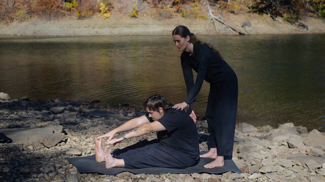 Outdoor yoga session with instructor assisting a seated forward bend by a riverside - Powered by Adobe