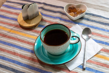Cup of black coffee with tea bags and sugar on striped tablecloth. Coffee break, morning ritual, home comfort. Real photo