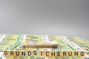 Wooden letter tiles spell Grundsicherung in front of stacked euro banknotes. Minimal grey background with copy space, symbolizing social benefits and income support.