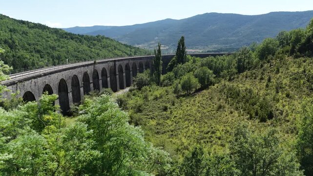 Curving railway viaduct arching through verdant mountain valley, showcasing scenic transportation infrastructure amid lush green landscape under bright summer sunlight