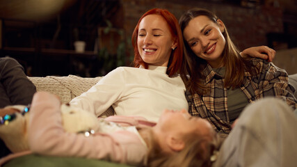 Two mothers laughing together while daughter rests on their laps during cozy family evening. Concept of family equality, modern parenting, diversity inclusion, and social and educational content.