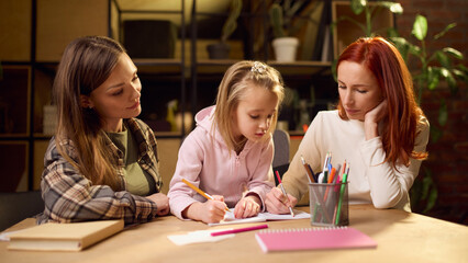 Two women helping little girl with drawing at home table. Concept of education, shared learning,...