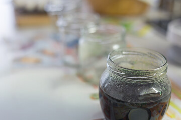 A row of jars with a dark liquid, Easter egg dye, on a light table. The foreground focuses on one jar, which is dewy. The blurred background suggests home preparation for Easter