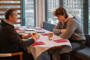 Young caucasian man and older man sitting at table, drinking coffee and talking in relaxed urban interior cafe with large windows. Meeting father and son. Two men talking in restaurant