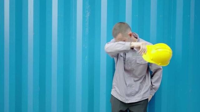 A worker work hard at container yard. Tired male engineer wipes sweat from brow, standing in front of warehouse logistic in cargo freight ship, Heavy profession. Physical work in hot weather.