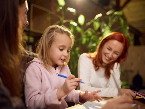Little girl drawing under warm light with two mums beside her. Concept of nurturing creativity, supportive home learning, inclusive family scenes, and emotional bonding. - Powered by Adobe