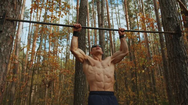 Shirtless athlete training in an autumn forest on makeshift sports ground and performs pull-ups on horizontal bar nailed to trees. Tracking shooting.