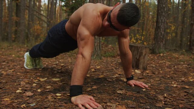 Shirtless bodybuilder does push-ups on makeshift sports ground in an autumn forest and gets up to take break after exercise.