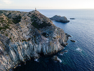 Aerial view of the rugged coastline cliffs meeting the deep blue sea, topped with a lighthouse standing tall against the horizon, Menorca, Spain.