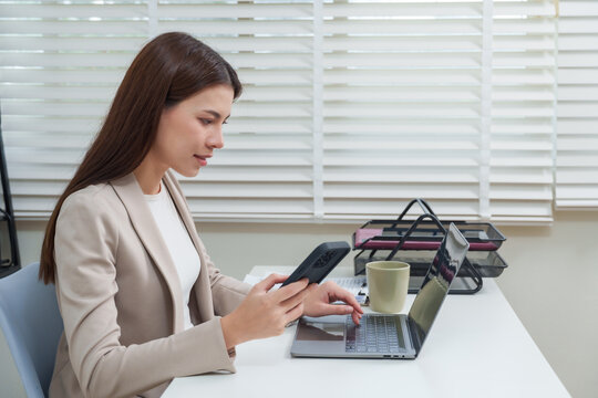 Asian businesswoman sitting at desk using laptop and smartphone together performing multitasking communication representing modern corporate lifestyle technology connection and productive work