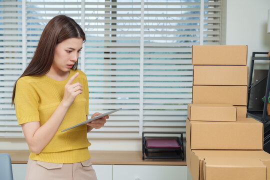 Asian woman using digital tablet to manage e-commerce sales and stock organization, surrounded by cardboard boxes, concept of online business productivity and small enterprise management