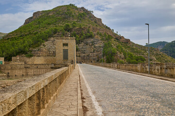 Fototapeta premium Scenic road built along the top of a massive stone dam, surrounded by mountain hills and greenery in Spain.