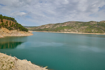 Calm turquoise reservoir surrounded by rocky hills and green slopes under cloudy sky. Serene...