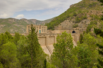 Straight road crossing a historic stone dam structure with mountains and dense forest surrounding the valley.