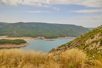 Obraz premium Wide reservoir with turquoise water, rocky shoreline and green pine-covered hills under a cloudy summer sky.