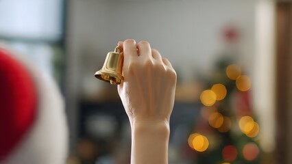 A hand wearing part of a Santa hat, holds a golden Christmas bell, ready to ring with blurred festive bokeh lights in the background, signaling holiday cheer.