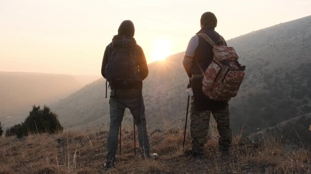 Back view of two men with backpacks standing in the mountains facing the sunset and watching the landscape while traveling together.