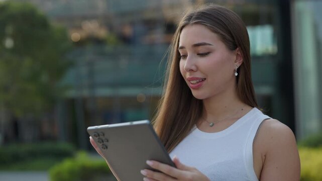 Portrait of pretty happy woman watches video via digital tablet on city street. Smiling female with long loose hair holds modern portable computer standing in summer urban garden
