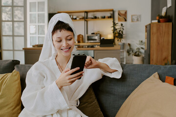 Young adult Caucasian woman sitting on sofa smiling while using smartphone for online shopping in modern home living room, wearing bathrobe and towel on head