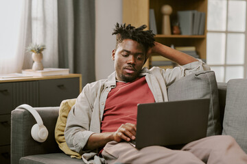 Young adult Black man sitting on sofa browsing laptop with relaxed posture, engaging in online shopping at home, wireless headphones resting nearby, focused on screen