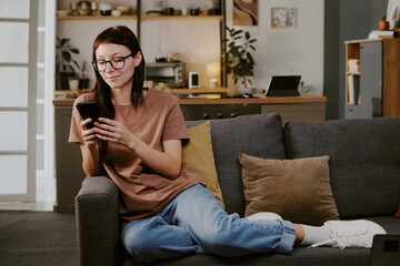 Obraz premium Caucasian young adult woman sitting on sofa using smartphone for online shopping, smiling while browsing digital store in modern living room, relaxed posture, eyeglasses visible