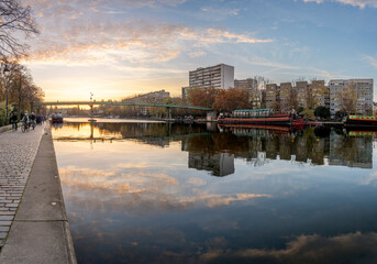 Naklejka premium Paris, France - 11 08 2025: Ourcq Canal. Panoramic View of the Villette Basin, Moselle Footbridge and buildings behind reflecting on the water at sunset and golden hour